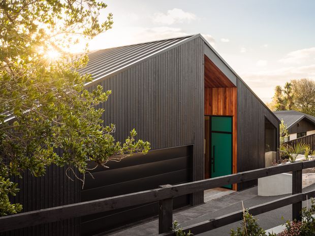 Exterior view of a contemporary gable-roofed residence with dark vertical cladding, a contrasting timber-lined recessed entry, and a vivid green front door. A dark garage door sits beneath the main volume, with a paved approach and timber boundary fence in the foreground. The house is set among established vegetation in warm late-day light.