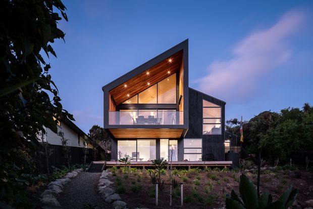 Twilight exterior view of a contemporary multi-level house with a dramatic angular gabled roofline, dark vertical cladding, extensive glazing, and a warm timber-lined recessed upper facade with integrated spotlights. A projecting upper-level balcony overlooks a landscaped front garden and gravel path.