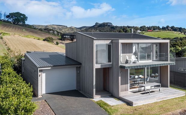 Exterior view of a compact contemporary two-storey house on a sloping rural/suburban site. The home has dark vertical cladding, a low-pitched metal roof, extensive glazed openings, a projecting upper-level balcony, an attached single-car garage, and a timber outdoor deck. Rolling landscaped terrain and neighbouring houses are visible in the background.