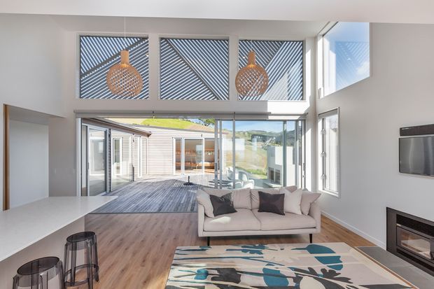 Bright contemporary open-plan living room with a double-height ceiling and extensive glazing opening onto a timber deck and landscaped courtyard. The room features pale timber flooring, white walls, a large light sofa, abstract patterned rug, suspended woven pendant lights, a wall-mounted television and a modern built-in fireplace. High clerestory windows include diagonal screening elements.