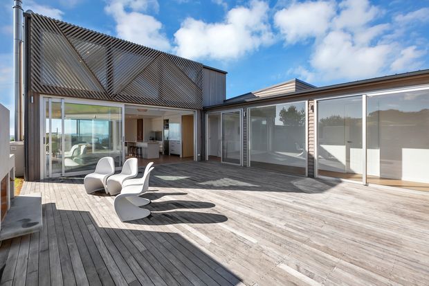 A spacious elevated timber deck courtyard adjoining a contemporary multi-wing house. The building features extensive floor-to-ceiling glazing, dark vertical and diagonal exterior screening, a low-pitched roof, and open-plan interiors visible through the sliding glass doors. Sculptural white outdoor lounge chairs sit on the weathered timber decking, with strong sunlight and long shadows creating a bright, relaxed outdoor setting.