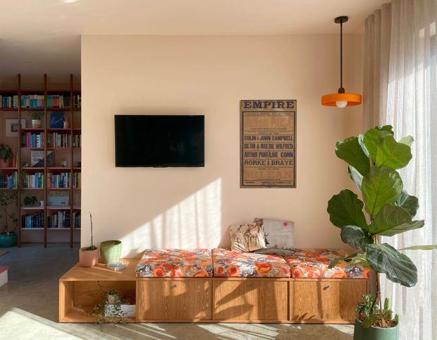 Warm, sunlit living room with a low timber built-in media console and daybed beneath a wall-mounted television. The room includes full-height sheer curtains, a large fiddle-leaf fig, open timber bookshelves filled with books, a vintage-style poster, and colourful patterned upholstery. Strong afternoon light creates a relaxed, eclectic atmosphere.
