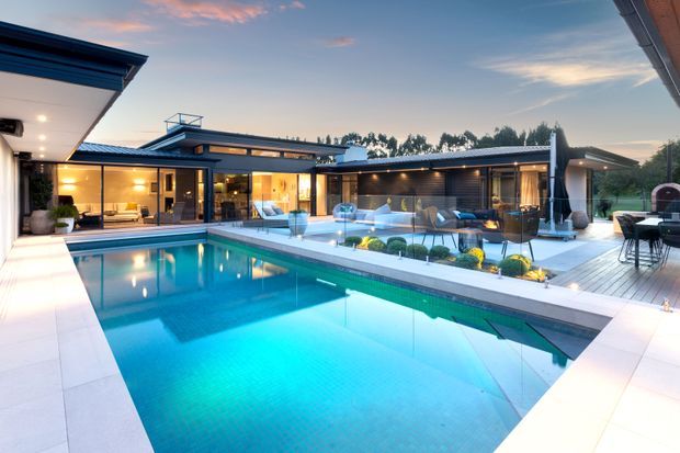 A contemporary single-storey home arranged around a large rectangular swimming pool and outdoor entertaining area. The image shows extensive floor-to-ceiling glazing, dark horizontal cladding, flat roof forms, a timber deck, outdoor seating, landscaped planting, and warm interior lighting at dusk.