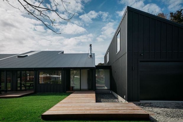 Exterior view of a contemporary dark-clad residential building composed of a low gabled-wing volume and a taller adjoining rectangular volume. The facade features vertical dark cladding, large glazed openings, a corrugated pitched roof with skylights, and a timber deck walkway leading across a lawn and gravel landscaping.