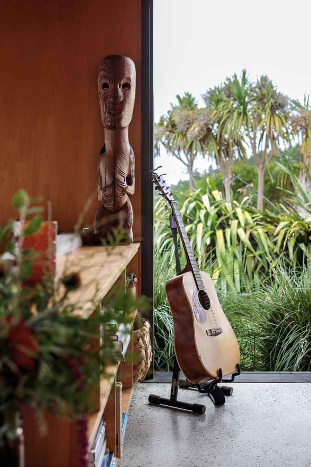 A warmly styled interior corner beside full-height glazing, featuring a carved wooden figurine displayed on open timber shelving and an acoustic guitar on a floor stand. Dense tropical planting is visible outside through the glazing. The space has a strong connection to nature, with timber joinery, a dark window frame, and a light speckled hard floor.