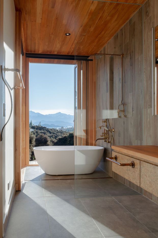 A luxurious contemporary bathroom with a freestanding white bathtub positioned beside full-height glazing overlooking a mountainous landscape. The room features large grey stone-look floor tiles, vertically grained stone-look wall cladding, warm timber lining across the ceiling and around the window, and exposed copper-toned plumbing with wall-mounted bath fittings. A floating timber-topped vanity extends along the right wall. Strong natural light creates a warm, refined atmosphere.
