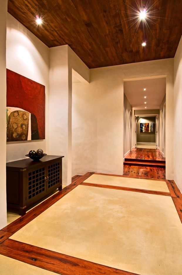 A warmly lit residential hallway with rich dark timber ceilings and hardwood floor inlays framing large cream-colored floor panels. White plaster walls, recessed downlights, a dark built-in console cabinet, and artwork create a refined, gallery-like transitional space leading to another corridor.