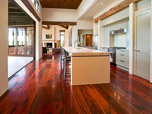 Open-plan kitchen and dining/living area with a large central island, white cabinetry, stainless-steel appliances, and a warm timber ceiling. Rich dark timber flooring extends through the space, with exposed timber beams and large glazed doors connecting to an outdoor area.