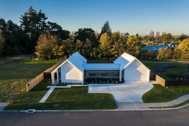 Aerial exterior view of a contemporary single-storey residential property with two white gabled wings connected by a lower central section, a pale blue-grey metal roof, timber privacy fencing, landscaped lawns, concrete driveway, and a suburban leafy setting.