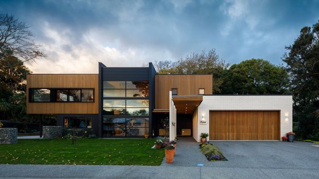 Street-facing exterior view of a contemporary multi-level residence with a strong geometric composition. The facade combines dark horizontal cladding, warm vertical timber-look wall sections, extensive floor-to-ceiling glazing, and a white masonry garage wing with a timber garage door. A landscaped front lawn, paved driveway, and planted entry path are visible, with mature trees surrounding the property.