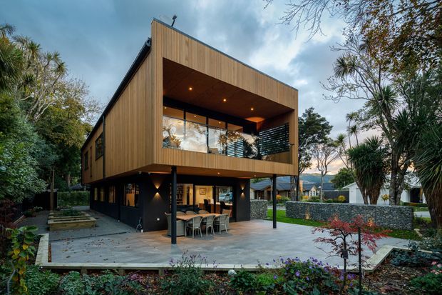 Exterior view of a contemporary two-storey home with a bold cantilevered upper volume clad in vertical timber battens. The lower level is dark and recessed, opening onto a paved outdoor dining and entertaining area, while the upper floor features a deep covered balcony with recessed downlights and extensive glazing. Landscaped gardens, stone retaining walls, and mature vegetation surround the building.