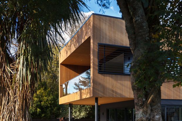 Exterior view of a contemporary elevated two-storey building surrounded by mature vegetation. The facade is clad in warm vertical timber boards, with a projecting upper volume, recessed balcony or terrace, large reflective glazing, and dark-framed louvred windows. Slim structural posts lift part of the building above the ground, creating a strong cantilevered form.