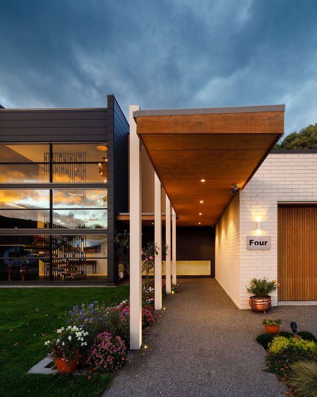 A contemporary residential entrance viewed from the exterior at dusk. The facade combines a dark horizontal-clad volume with extensive floor-to-ceiling glazing and a visible stair, alongside a white brick volume with a tall timber entry door. A broad timber-lined canopy projects over the covered walkway, supported by slender light-coloured posts. Recessed canopy lighting, landscaped garden beds, potted plants, and the house number 'Four' are visible.