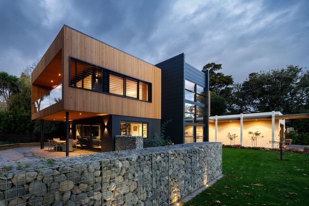 A contemporary multi-level residence photographed at dusk, featuring a cantilevered upper volume clad in vertical timber, dark metal architectural elements, expansive horizontal windows with external louvres, and a glazed stair or circulation volume. A covered outdoor terrace and a long low ancillary wing extend across the landscaped garden. The foreground is defined by a substantial gabion stone retaining wall, with warm interior and exterior lighting contrasting against the cool overcast sky.