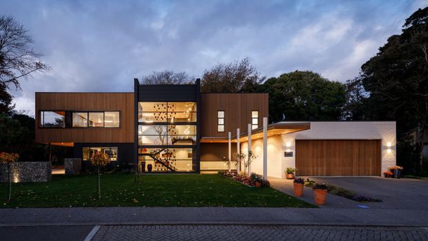Contemporary multi-level residence photographed at dusk, featuring a dark timber-clad rectangular wing, a prominent double-height glazed central stairwell, and a connected single-storey garage volume with a broad cantilevered roof. The house sits in a landscaped suburban setting with a paved driveway and lawn.