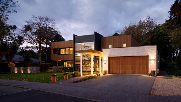 Twilight exterior photograph of a contemporary multi-level residence. The home features a combination of dark vertical cladding, warm timber wall cladding, extensive glazing, a projecting entry canopy, and a pale brick-clad garage wing with a timber double garage door. A landscaped front lawn, driveway, and mature trees frame the facade.