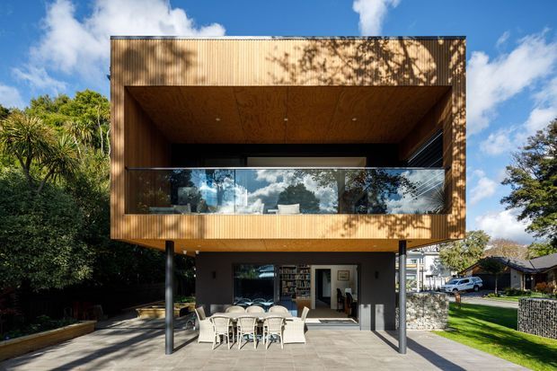Front exterior view of a contemporary elevated house with a box-like upper storey clad in warm vertical timber, a deeply recessed balcony with clear glass balustrading, and a covered ground-level outdoor dining area supported by slender dark columns. The home sits in a leafy suburban setting with strong daylight and a blue sky.