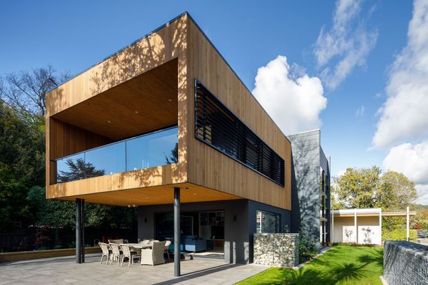 A contemporary multi-level house viewed from the exterior, featuring a strong rectangular timber-clad upper volume, deep covered balcony, glass balustrades, dark horizontal louvres, slender steel columns, and a partially open ground-level outdoor living area. Stonework forms the lower wall, with a lawn and landscaped suburban setting visible around the building.