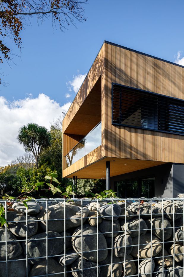 Exterior view of a contemporary multi-level house with a projecting upper storey clad in vertical warm timber boards. The design features a deep cantilevered overhang, large glazed openings, dark horizontal window louvres, slender dark structural supports, and a gabion stone retaining wall in the foreground. The house is set among established vegetation under a clear blue sky.