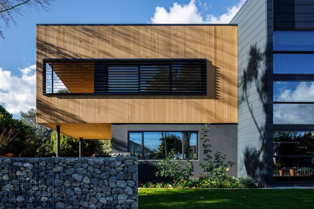 Exterior view of a contemporary multi-level residential building with a prominent upper box clad in warm vertical timber slats, dark horizontal louvres, and a projecting cantilever. The lower level combines dark grey walling, glazing, and a gabion stone boundary wall, while an adjacent section features grey horizontal cladding and large windows. Landscaped garden and blue sky surround the building.