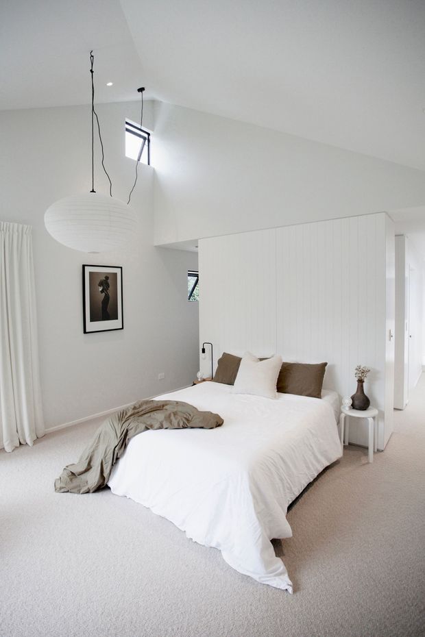 A bright, minimalist bedroom with a low-profile bed dressed in white bedding and taupe cushions, positioned against a white vertical-panel feature wall. Soft white curtains, pale carpet, a small round bedside table, framed artwork, and a large paper pendant light create a calm, neutral interior. A partially open doorway and high windows are visible in the background.