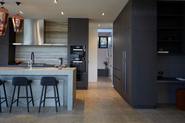 Contemporary open-plan kitchen with a large pale stone island and breakfast bar, dark timber cabinetry, integrated double ovens, stainless-steel rangehood, and a horizontally textured stone splashback. The kitchen opens toward a hallway and includes a dark built-in shelving area on the right. Recessed ceiling lighting, pendant lights, and warm neutral flooring create a refined, modern atmosphere.