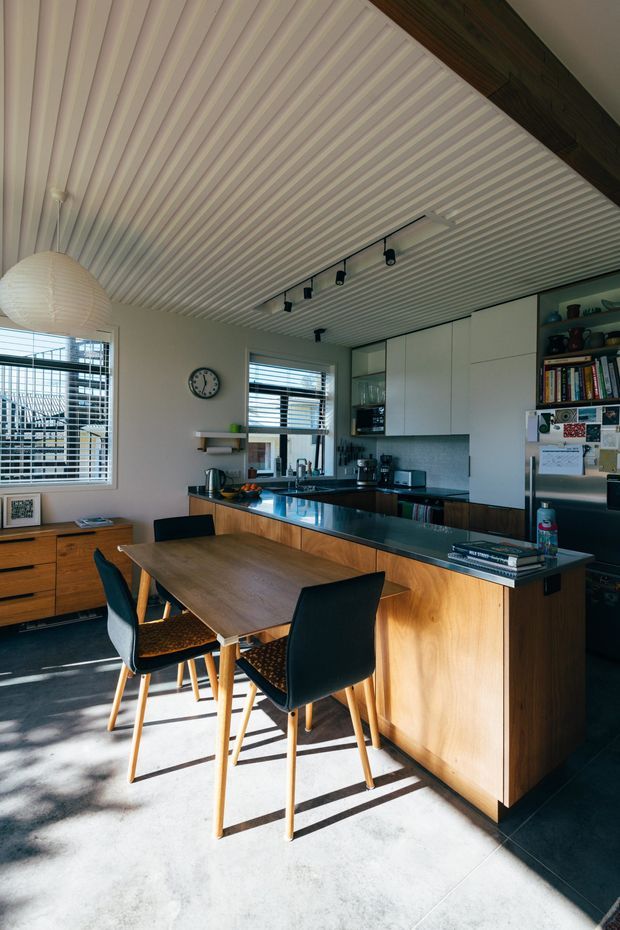 Bright open-plan kitchen and dining area with a timber island and dining table, black upholstered timber-leg chairs, dark benchtops, white cabinetry, tiled flooring, and a distinctive ribbed white ceiling with track lighting. Large louvred windows bring in strong natural light, while open shelving and a refrigerator add lived-in detail.