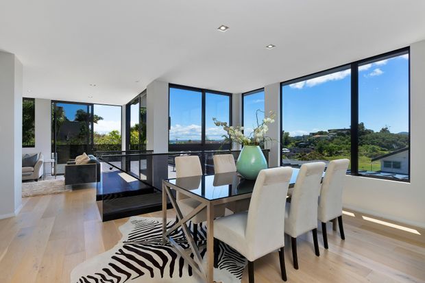Bright contemporary dining area with a large rectangular dining table and upholstered chairs, positioned beside expansive black-framed corner windows. Pale timber flooring, white walls and recessed ceiling lights create a clean, airy interior, while the glazing overlooks a leafy suburban landscape and water or sky beyond. A zebra-pattern rug and large turquoise vase provide accents.