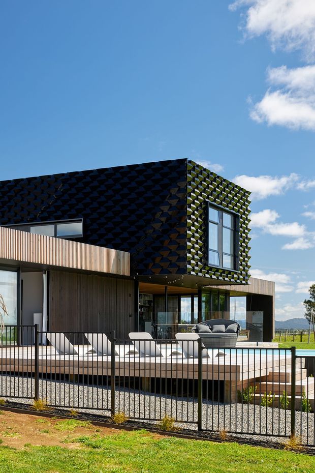 A contemporary two-storey house viewed from the front garden, featuring a dark geometric patterned upper facade, timber-clad lower level, expansive glazing, and a projecting balcony or terrace. A landscaped pool deck with outdoor seating and a black metal fence occupies the foreground, with open rural landscape and blue sky beyond.