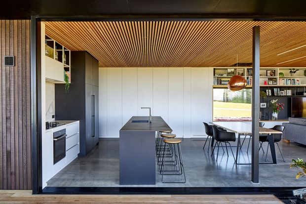 Open-plan contemporary kitchen and dining area viewed through large sliding glass doors. The space features a dark central island, white handleless cabinetry, integrated appliances, a grey concrete-like floor, a warm slatted timber ceiling, dark vertical timber wall cladding, black-framed glazing, a dining table with black chairs, open shelving, and a warm copper pendant light.