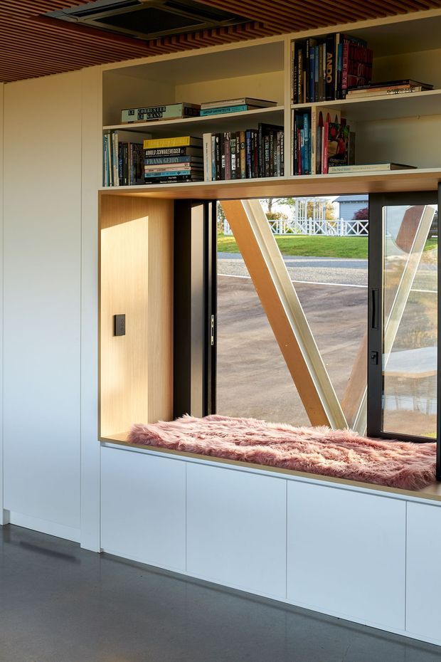 A bright, compact interior featuring full-height white built-in cabinetry, open timber-lined shelving filled with books, and a deep window seat beneath a large glazed opening. The window seat has a soft pink faux-fur cushion, while exposed angled timber structure is visible outside. A slatted timber ceiling and polished grey floor add warmth and contrast to the clean built-in joinery.