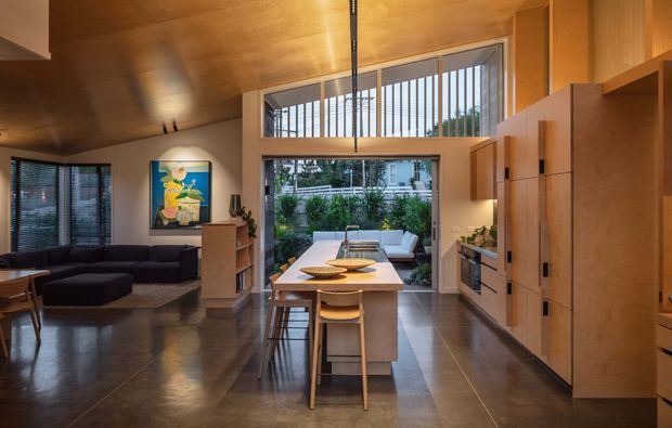 A warm, open-plan contemporary living and dining area with a compact kitchen and central island. The room features extensive timber lining on the ceiling and built-in cabinetry, polished dark flooring, a large glazed opening to a planted outdoor area, high clerestory windows with vertical screening, and a dark sectional lounge. A dining table and stools occupy the island, while the kitchen joinery runs along the right wall.