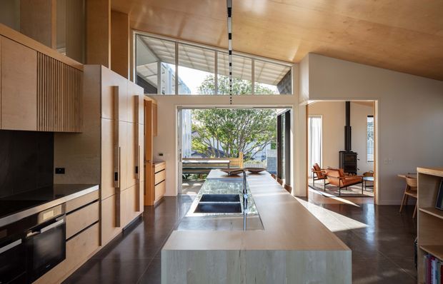A spacious open-plan kitchen with a large central island, full-height glazing and a timber-lined ceiling. The kitchen includes tall light-wood cabinetry, integrated dark appliances, a pale stone island benchtop and dark flooring. Beyond the kitchen is a bright living area with a freestanding wood-burning stove, lounge seating and outdoor views through large glazed doors.
