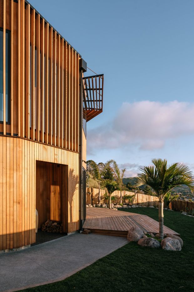 Exterior view of a contemporary timber-clad building with strong vertical wood battens, a recessed timber entry door, and a projecting upper balcony with timber railing. A timber deck, paved path, lawn, ornamental palms, boulders, and a timber boundary fence surround the building, with hills visible in the distance.