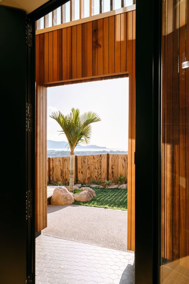 View from inside through a tall open timber-framed doorway toward a landscaped coastal courtyard. The opening is clad in warm vertical timber boards with high clerestory glazing above, dark-framed doors at the sides, a white hexagonal-tile threshold, and an exterior area with a palm, timber fence, rocks, lawn, and ocean horizon beyond.