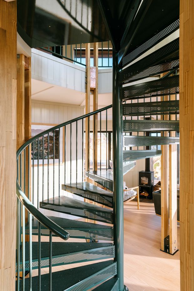 Interior view of a dramatic multi-level stairwell featuring a perforated black metal spiral staircase with curved railing and slender balusters. The stair is set within a light-filled open-plan timber-lined interior, with exposed vertical timber posts, pale panelled walls, expansive glazing, and a glimpse of a living area with a wood-burning stove below.