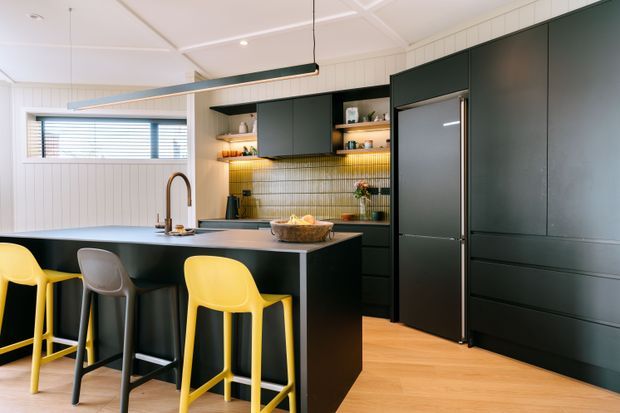 A bright, contemporary kitchen with a large dark island, integrated dark cabinetry and refrigerator, open shelving, and a warm illuminated tiled backsplash. Yellow and grey stools provide colourful accents at the island. White vertical wall panelling, timber flooring, and a vaulted panelled ceiling create a clean yet warm interior.