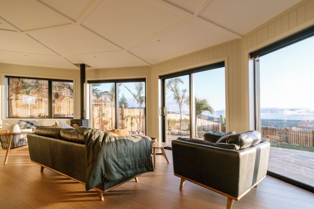 Bright coastal living room with expansive corner glazing and sliding doors opening toward a timber deck and ocean view. The room features warm timber flooring, cream vertical wall panelling, a light coffered ceiling, a large dark leather sectional sofa, a matching armchair, and a compact dining area. Palm trees and the waterfront are visible outside.
