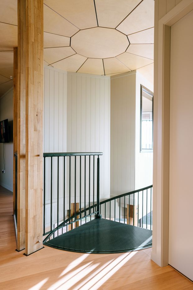 Bright interior view of a stair landing with a distinctive circular/spiral stair descending through an opening. The space features pale timber flooring, vertical white wall panelling, a sculptural segmented timber ceiling, exposed timber posts, and dark green metal balustrades. Sunlight casts strong linear shadows across the floor.