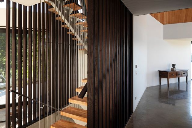 A contemporary interior entry and stairway featuring a sculptural timber staircase enclosed by a tall dark timber vertical-slat screen. Natural light filters through the slats from glazing on the left, while a polished concrete floor, white walls, timber ceiling accents, and a console table define the adjacent open hallway. The image highlights the stair structure, timber detailing, and open-plan circulation space.