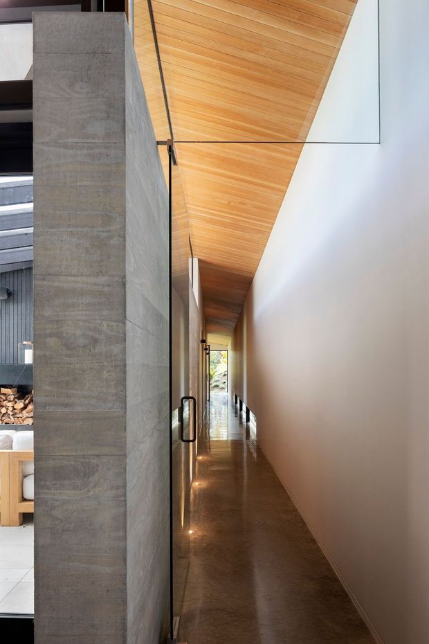 A narrow, elongated interior hallway beside a tall exposed concrete wall, with a polished concrete floor, warm timber-lined angled ceiling, smooth white wall, and black-framed glass doors. Recessed floor lights lead toward a bright glazed opening at the far end. A partial living area with firewood and furnishings is visible through the glass on the left.