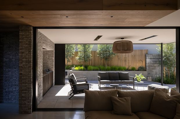 A warm, contemporary indoor-outdoor living area opening onto a landscaped courtyard. The interior features a large brown sectional sofa, black lounge chairs, a low coffee table, exposed brick walls, dark-framed full-height glazing, timber ceiling lining, recessed linear lighting, and a woven pendant. Outside is a paved patio with garden planting, timber fencing, and masonry boundary walls.