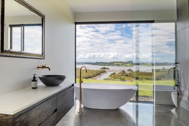 Contemporary bathroom with a freestanding oval bathtub positioned beside floor-to-ceiling glazing overlooking a coastal or estuarine landscape. The room includes a dark vessel basin on a white vanity with timber-toned drawer fronts, a wall-mounted brass tap, polished grey floor, and a large framed mirror. A separate toilet and towel rail are visible at the right.