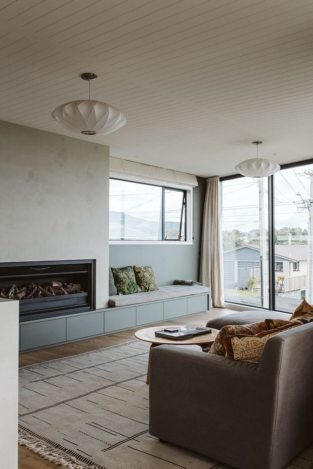 A warm, contemporary living room with a built-in linear fireplace and low blue-grey cabinetry beneath a window seat. The room has pale timber flooring, a large neutral patterned rug, a grey upholstered sofa and armchairs, sculptural pendant lights, and extensive windows and glazed doors opening to an exterior view. The ceiling is clad in narrow white-painted boards, and curtains frame the glazing.