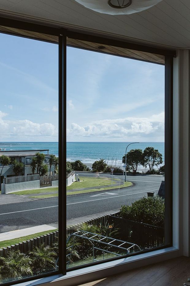 Interior view through large floor-to-ceiling windows overlooking a coastal residential street and turquoise ocean. The room has dark window frames, light wood flooring, a white paneled ceiling, and a simple circular ceiling light. The outlook is bright and open, with landscaped gardens, roadways, and beachfront visible beyond the glazing.