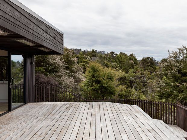 A contemporary timber-clad building opens onto a large elevated timber deck with dark metal balustrading. The deck overlooks dense native bushland beneath an overcast sky. A glazed doorway and covered roof structure are visible at the left edge.