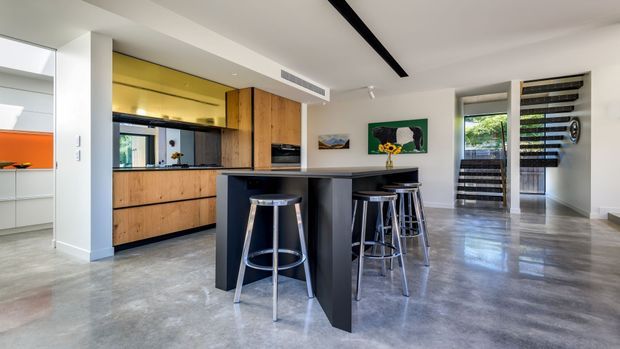 Open-plan contemporary kitchen and dining area with a dark island and bar seating, warm timber cabinetry, polished concrete flooring, white walls and ceiling, and a large glazed opening with horizontal external screening. A bright yellow glass backsplash or feature panel adds colour behind the kitchen.