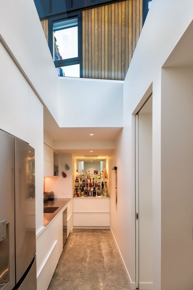 A narrow, bright interior passage combining a compact galley kitchen with a built-in drinks bar at the far end. White cabinetry and walls frame a polished concrete-toned floor, while a double-height void and skylight-like upper glazing bring in natural light. Vertical timber wall panelling and dark-framed windows add warmth and contrast.
