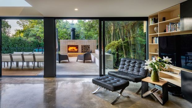 Contemporary living room with polished concrete flooring, expansive black-framed glazing opening to a leafy outdoor terrace, and a built-in timber shelving and media wall. A black leather lounge chair and ottoman sit beside a small timber side table, while an outdoor fireplace is visible beyond the open glazing.