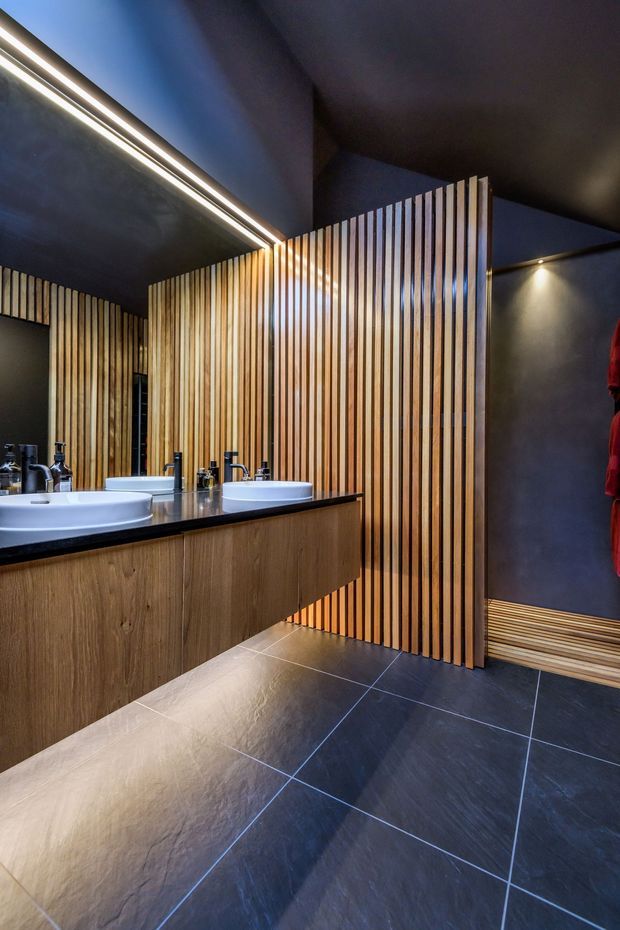 A dark, contemporary bathroom featuring a long floating timber vanity with a black stone benchtop, multiple white vessel basins, and a large mirror with integrated linear lighting. Vertical timber slat screening forms a partition and feature wall, while large dark floor tiles, charcoal walls, and recessed spotlights create a dramatic, refined atmosphere.